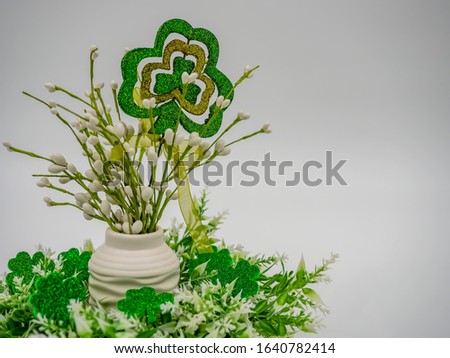 White beaded flower arrangement surrounded by greenery and glittery four leaf clovers with a big four leaf clover and a green bow on a white background for St. Patrick’s Day!