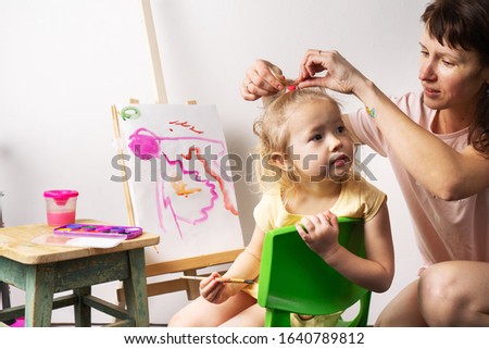 Children's creativity: a little girl draws with a brush and watercolors on canvas with an easel. Mom straightens the hair of the child.