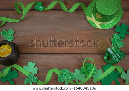 Saint Patrick's Day image of glittered green and felt shamrocks with ribbon form a border on a rustic wooden background. Leprechaun hat and pot of gold included. Green glitter scattered around.