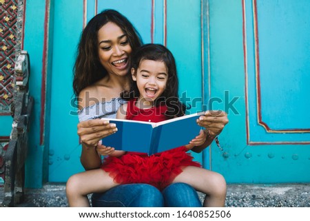 Asian young beautiful lady and cute preschool daughter reading book with blue cover and sitting on stairs near doors while laughing
