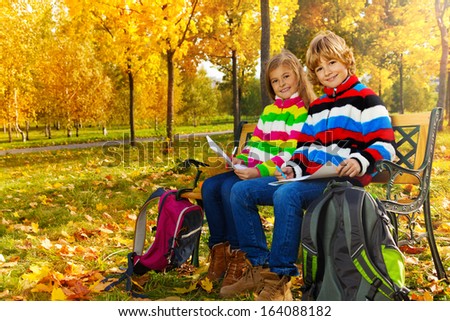 Portrait of two happy blond kids, twins sitting on the bench after school drawing pictures on paper with backpacks 