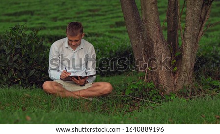 man sits under a tree on the grass and writes in a notebook.