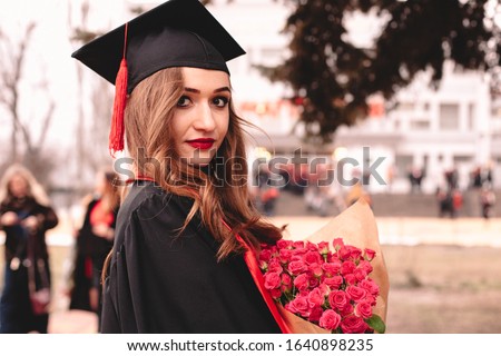Portrait of young female graduate student in mortarboard holding bouquet of flowers while standing in campus at graduation ceremony Royalty-Free Stock Photo #1640898235