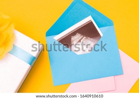 a picture of an ultrasound in a blue envelope,a silver box with a blue ribbon and a yellow flower and a pink envelope made of fabric, yellow background copy space top view, woman pregnant concept