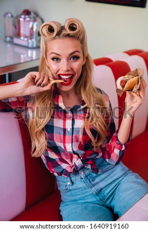 Photo of charming joyful woman with beautiful hairstyle eating hotdog and french fries in retro american cafe