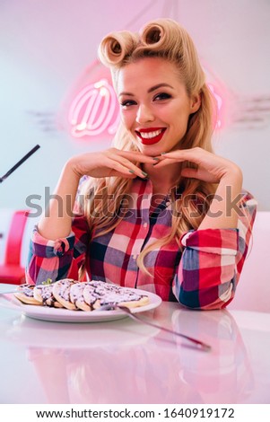 Photo of gorgeous cheerful woman with beautiful hairstyle eating pancakes and smiling in retro american cafe