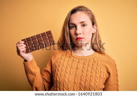 Young beautiful blonde woman holding chocolate bar standing over isolated yellow background with a confident expression on smart face thinking serious