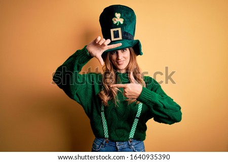 Beautiful brunette woman wearing green hat with clover celebrating saint patricks day smiling making frame with hands and fingers with happy face. Creativity and photography concept.