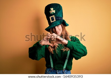 Beautiful brunette woman wearing green hat with clover celebrating saint patricks day smiling in love doing heart symbol shape with hands. Romantic concept.
