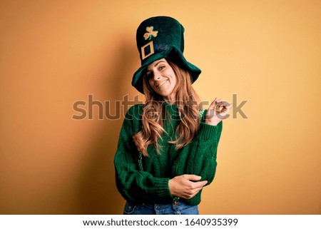 Beautiful brunette woman wearing green hat with clover celebrating saint patricks day with a big smile on face, pointing with hand and finger to the side looking at the camera.