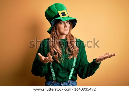 Beautiful brunette woman wearing green hat with clover celebrating saint patricks day clueless and confused expression with arms and hands raised. Doubt concept.