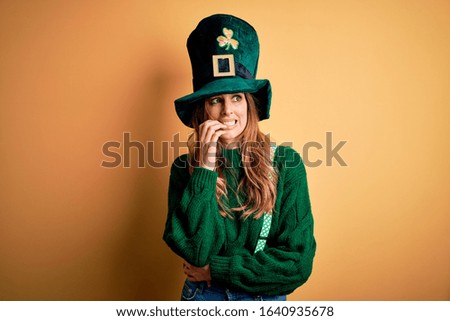 Beautiful brunette woman wearing green hat with clover celebrating saint patricks day looking stressed and nervous with hands on mouth biting nails. Anxiety problem.