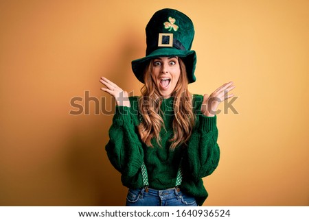 Beautiful brunette woman wearing green hat with clover celebrating saint patricks day celebrating crazy and amazed for success with arms raised and open eyes screaming excited. Winner concept