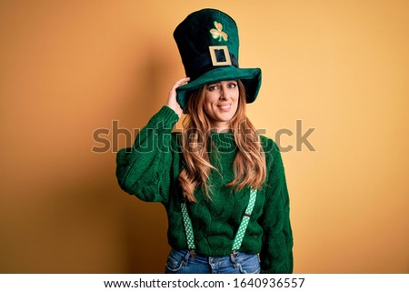 Beautiful brunette woman wearing green hat with clover celebrating saint patricks day confuse and wonder about question. Uncertain with doubt, thinking with hand on head. Pensive concept.