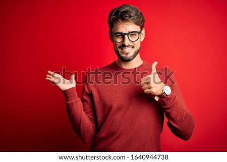 Young handsome man with beard wearing glasses and sweater standing over red background Showing palm hand and doing ok gesture with thumbs up, smiling happy and cheerful