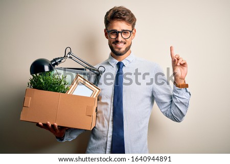 Young businessman with beard fired holding cardboard standing over white background surprised with an idea or question pointing finger with happy face, number one