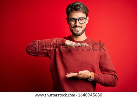 Young handsome man with beard wearing glasses and sweater standing over red background gesturing with hands showing big and large size sign, measure symbol. Smiling looking at the camera. Measuring 
