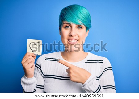 Young woman with blue fashion hair holding romantic heart shape on note over isolated background very happy pointing with hand and finger