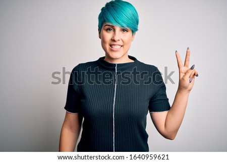 Young beautiful woman with blue fashion hair wearing casual t-shirt over white background smiling with happy face winking at the camera doing victory sign. Number two.