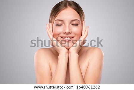Optimistic young woman with closed eyes enjoying softness of perfect skin against gray background