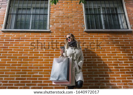 Blonde girl with white coat and black glasses is leaning on a brick wall with bags and a cup of coffee