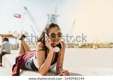 A smiling woman lies by the beach. She wears sunglasses and talks on cell phone