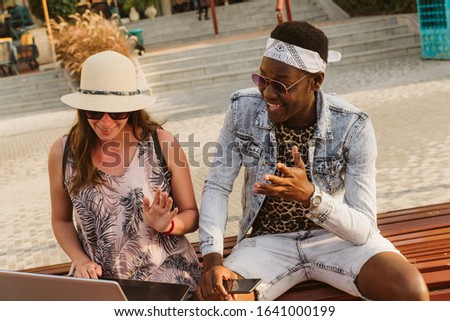A smiling woman with a hat and sunglasses holds a laptop in her lap. Next to her is an African-American who is laughing