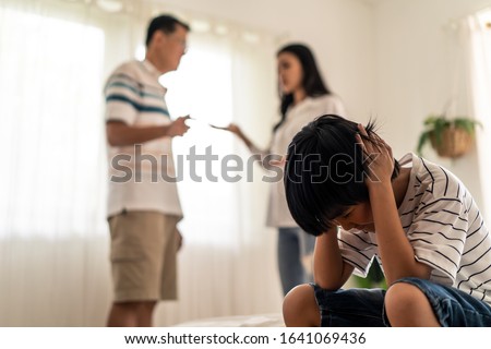 Asian boy kid sitting and crying on bed while parents having fighting or quarrel conflict at home. Child covering face and eyes with hands do not want to see the violence. Domestic problem in family. Royalty-Free Stock Photo #1641069436