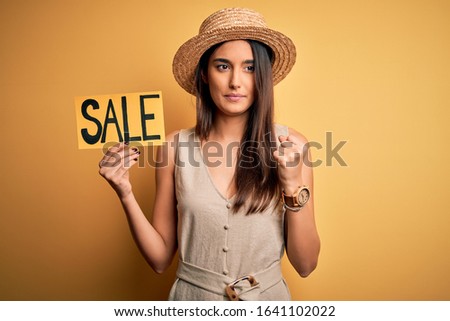 Young beautiful brunette woman wearing hat holding sale banner over yellow background pointing with finger to the camera and to you, hand sign, positive and confident gesture from the front