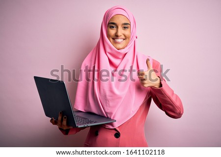 Young beautiful businesswoman wearing pink muslim hijab and business jacket using laptop happy with big smile doing ok sign, thumb up with fingers, excellent sign