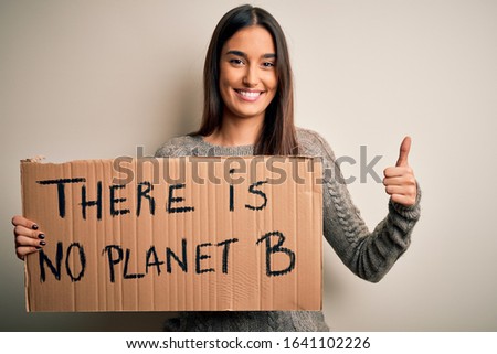 Young beautiful brunette activist woman protesting for save the planet holding banner happy with big smile doing ok sign, thumb up with fingers, excellent sign