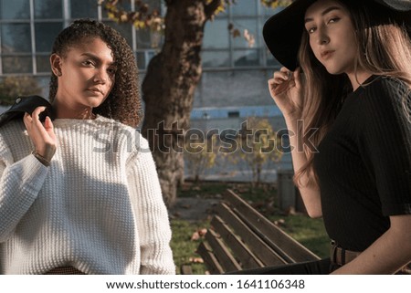 pair of young beautiful mexican generation y girls. The girl is sitting on the back of the bench, a girlfriend is standing next to curly hair and a white sweater, holding a jacket on her shoulder