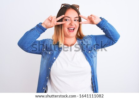 Beautiful woman wearing denim shirt standing over isolated white background Doing peace symbol with fingers over face, smiling cheerful showing victory