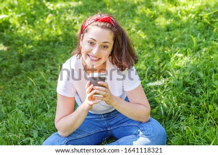 Happy girl smiling outdoor having lunch break. Beautiful young brunete woman with take away coffee cup resting on park or garden green grass background. Education freelancer resting leisure concept