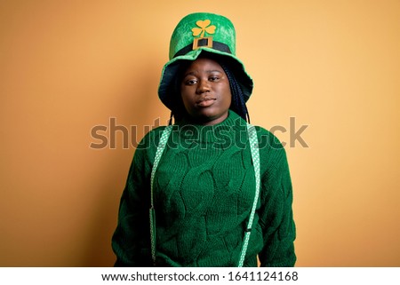 Plus size african american woman with braids wearing green hat with clover on st patricks day Relaxed with serious expression on face. Simple and natural looking at the camera.