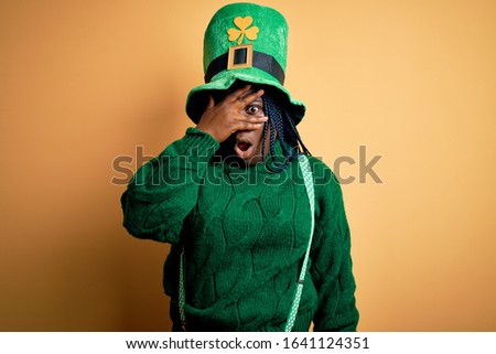 Plus size african american woman with braids wearing green hat with clover on st patricks day peeking in shock covering face and eyes with hand, looking through fingers with embarrassed expression.