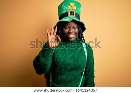Plus size african american woman with braids wearing green hat with clover on st patricks day smiling positive doing ok sign with hand and fingers. Successful expression.