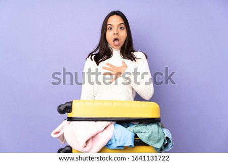 Traveler young woman with a suitcase full of clothes over isolated purple background surprised and shocked while looking right