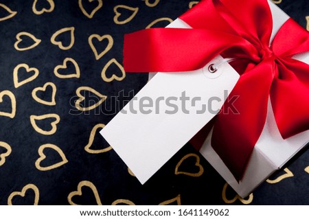 Blank gift tag on white Valentine's Day box with red bow sitting on a black background of golden hearts