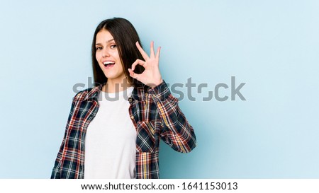 Young caucasian woman isolated on blue background winks an eye and holds an okay gesture with hand.