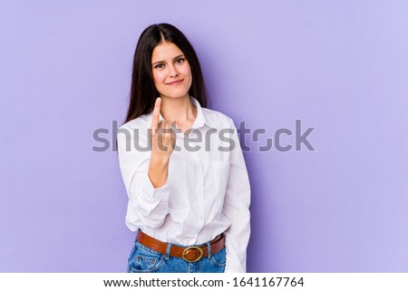Young caucasian woman isolated on purple background pointing with finger at you as if inviting come closer.