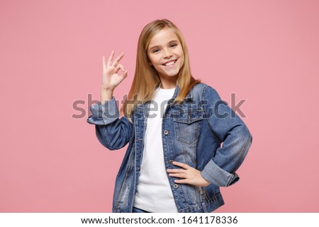 Smiling little blonde kid girl 12-13 years old in denim jacket posing isolated on pastel pink wall background children portrait. Childhood lifestyle concept. Mock up copy space. Showing OK gesture