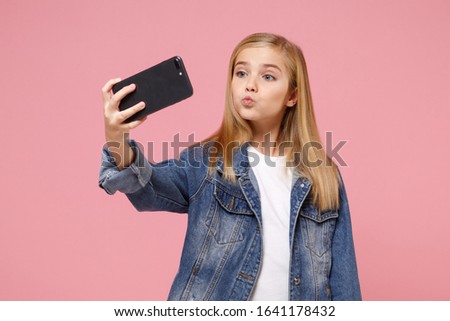 Pretty little blonde kid girl 12-13 years old in denim jacket isolated on pastel pink wall background. Childhood lifestyle concept. Mock up copy space. Doing selfie shot on mobile phone, blowing lips