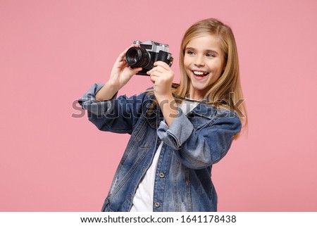 Cheerful little blonde kid girl 12-13 years old in denim jacket posing isolated on pastel pink background in studio. Childhood lifestyle concept. Mock up copy space. Hold retro vintage photo camera