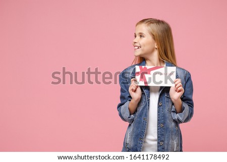 Smiling little blonde kid girl 12-13 years old in denim jacket posing isolated on pastel pink wall background. Childhood lifestyle concept. Mock up copy space. Hold gift certificate, looking aside