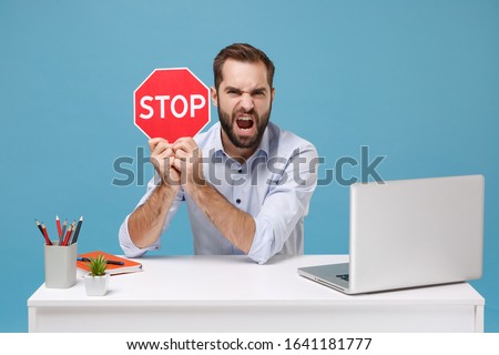 Angry irritated young man in light shirt work at white desk with pc laptop isolated on pastel blue background. Achievement business career concept. Mock up copy space. Hold red sign with Stop title