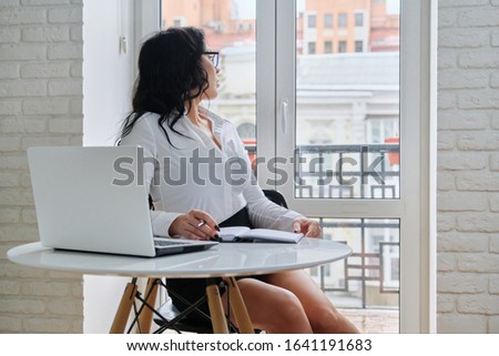 Beautiful business woman working with computer laptop, sitting at desk near window. Fashionable female in white shirt writing in business notebook