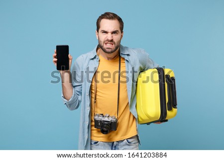 Irritated traveler tourist man in casual clothes with photo camera isolated on blue background. Passenger traveling abroad on weekends. Air flight journey Hold suitcase mobile phone with blank screen