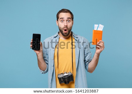 Shocked traveler tourist man in yellow clothes with photo camera isolated on blue background. Passenger traveling on weekends. Air flight journey. Hold passport tickets mobile phone with blank screen