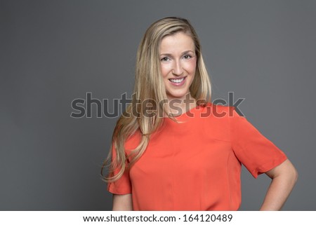 Stylish blond woman in a trendy orange top looking at the camera with a friendly smile against a grey studio background with copyspace
